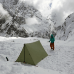 Green ultralight fly tent tarp setup on snow on a mountain, with a camper and mountains in the background.