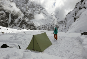 Green ultralight fly tent tarp setup on snow on a mountain, with a camper and mountains in the background.
